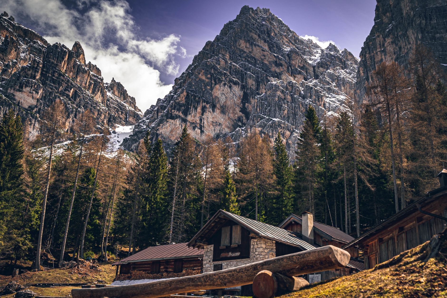 A scenic view of mountain cabins surrounded by towering mountains and trees under a cloudy sky.
