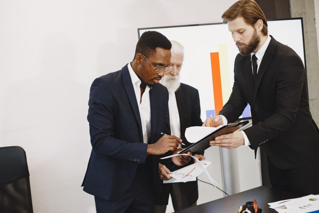 Three businessmen engaged in document signing in a modern office setting. — Take Profit Trader Allows You to Trade Without Us